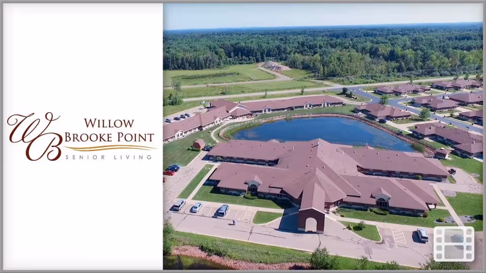 Aerial view of Willow Brooke Point Senior Living facility showing multiple connected buildings with brown roofs surrounding a central pond, with parking areas and green spaces, and a forested area in the background.