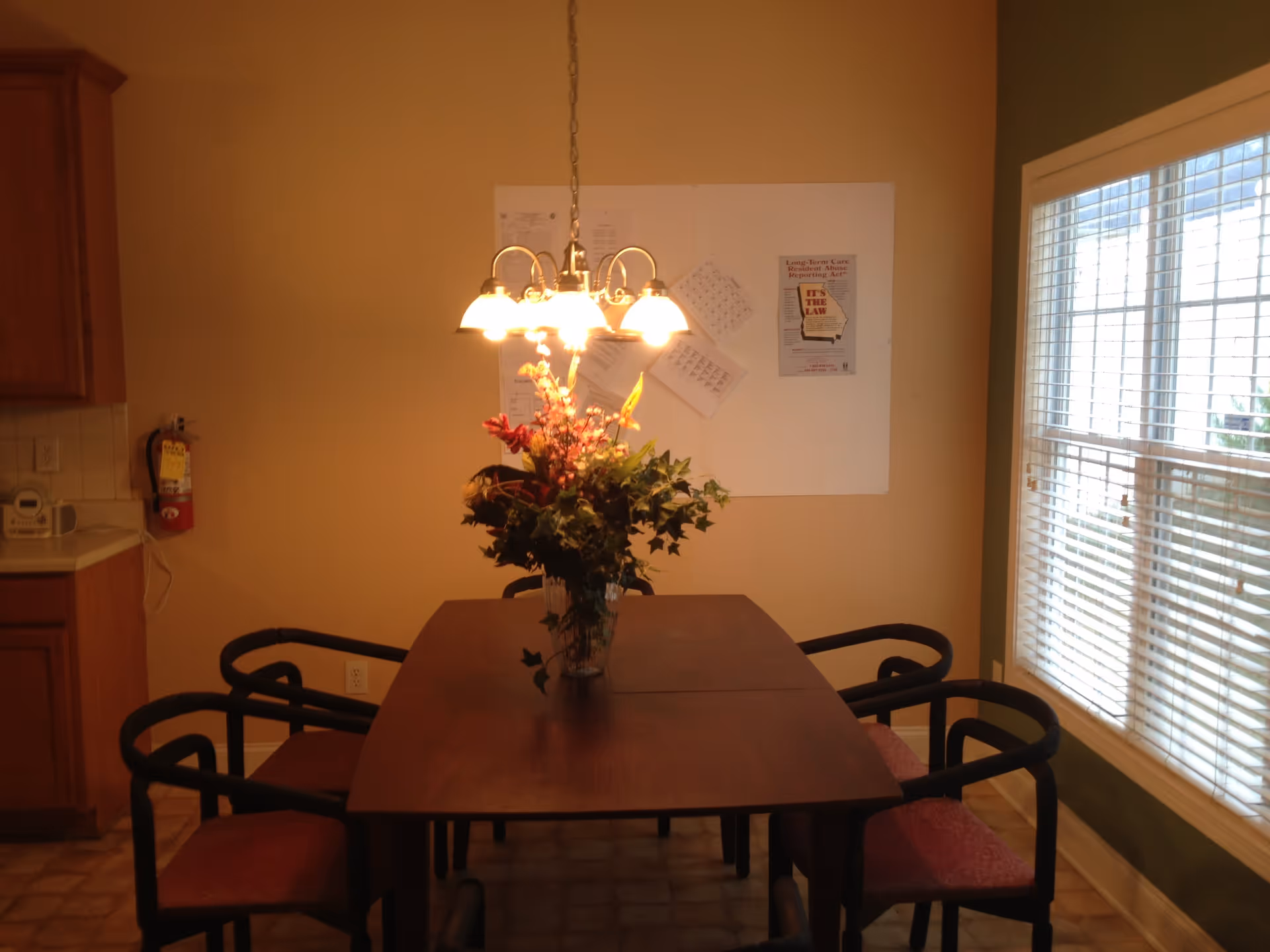 A dining area with a wooden table surrounded by six chairs with red cushions. A vase with a floral arrangement is placed in the center of the table. Above the table hangs a chandelier with five lit lamps. On the wall behind the table, there are several papers and a poster about Long-Term Care Resident Abuse Reporting Act. To the right, there is a window with white blinds letting in natural light. To the left, part of a kitchen counter and cabinets are visible along with a fire extinguisher mounted on the wall.