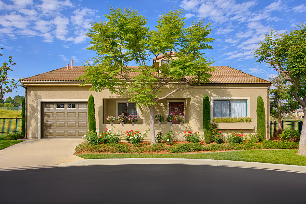 Front view of a single-story stucco house with a garage, landscaped flowerbeds, and a tree in the yard.