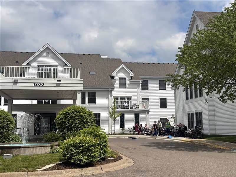 Exterior view of a white multi-story senior living facility building with a covered entrance labeled 1200. There is a fountain and green bushes in the foreground, and a group of people sitting and standing near the building on the right side. The sky is partly cloudy.