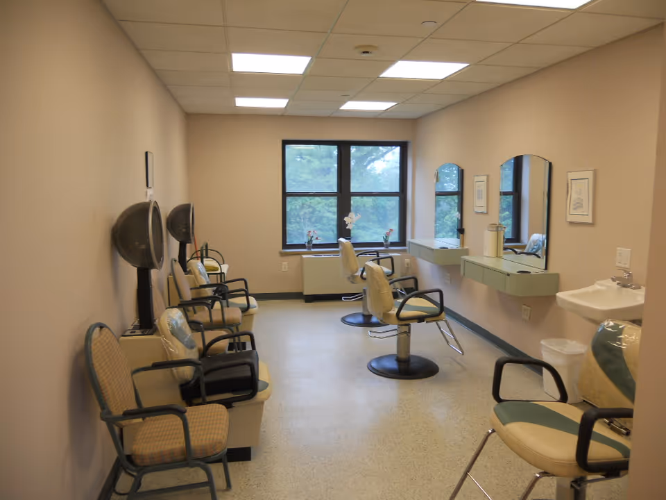 Small salon-style hairstyling room with multiple styling chairs, hooded dryers, mirrors, and a window at the far end.