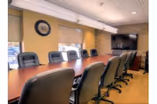 A conference room with a long wooden table surrounded by black office chairs on both sides. The room has beige walls, three windows with beige roller blinds, a wall clock, and a large flat-screen TV mounted on the wall at the far end.