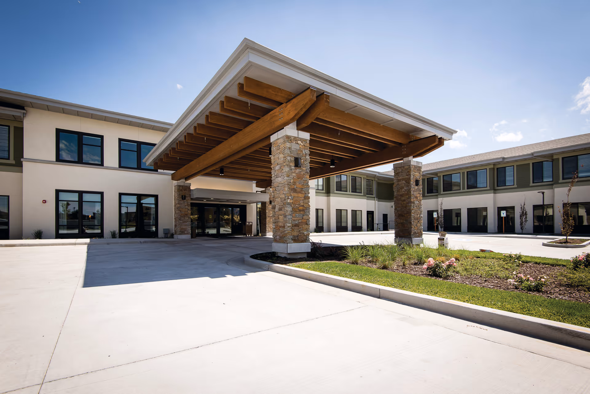 Exterior view of a modern senior living facility entrance with a large covered drop-off area supported by stone pillars and wooden beams. The building is two stories with multiple windows and landscaped greenery around the driveway.