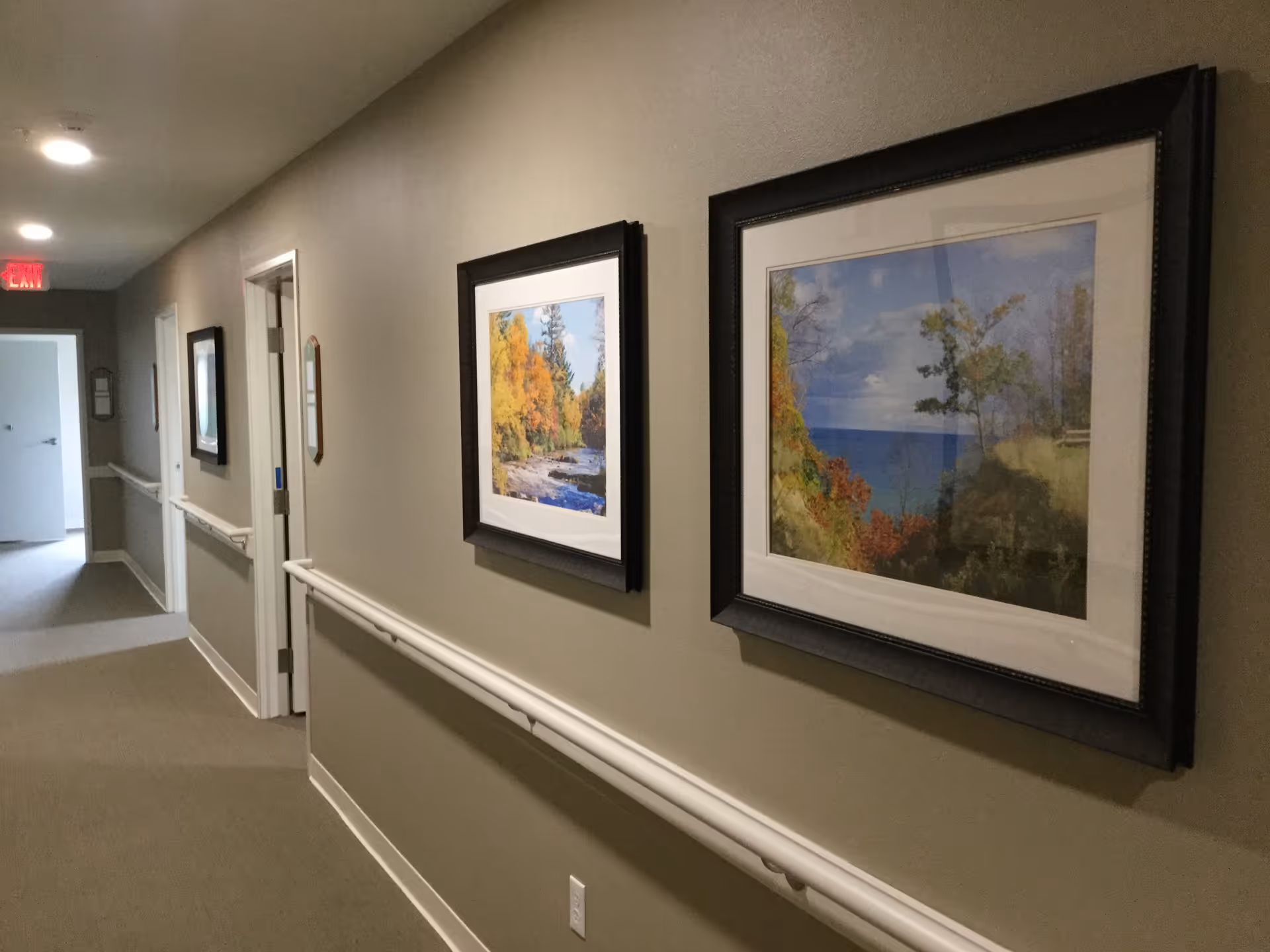 A well-lit hallway in a senior living facility with beige walls and carpeted floor. The hallway features handrails on both sides and framed landscape paintings hanging on the walls. Several doorways are visible along the corridor, and an exit sign is illuminated at the far end.