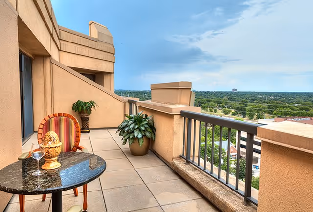 Balcony area with a small round table and a striped chair. On the table, there is a decorative item and a glass of water. Two potted plants are placed along the wall. The balcony overlooks a green landscape with trees and buildings under a partly cloudy sky.