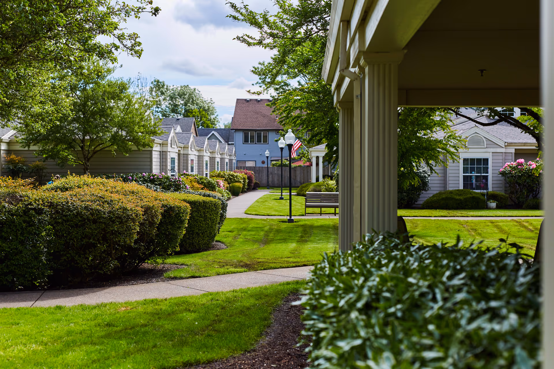 A well-maintained outdoor garden area at Jurgens Park Senior Living featuring green lawns, trimmed bushes, trees, a paved walkway, benches, and several small residential-style buildings with light-colored siding and windows. The sky is partly cloudy.
