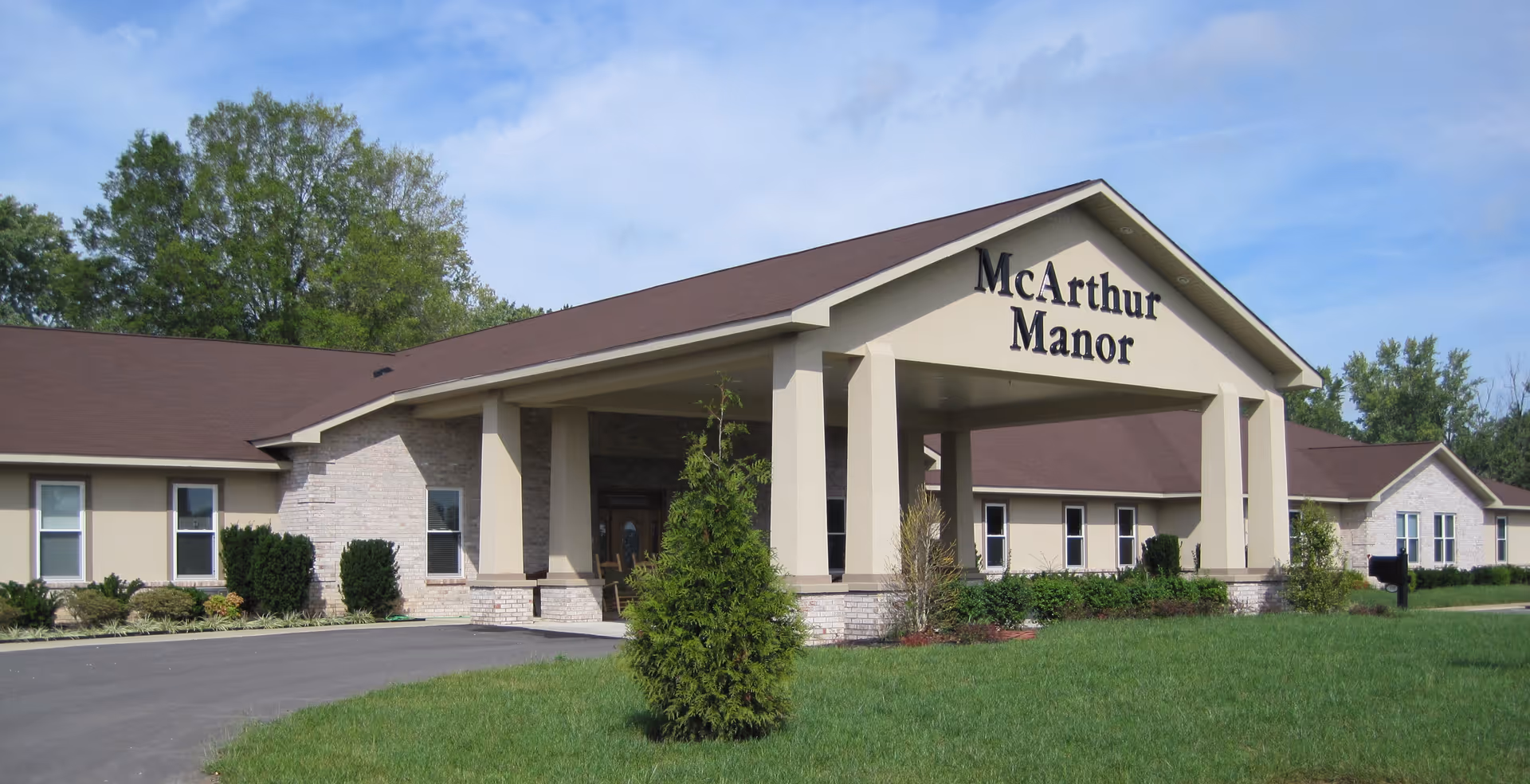 Front entrance of McArthur Manor assisted living building with a covered portico, columns, and lawn.