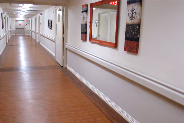 A long, well-lit interior hallway in a nursing and rehabilitation center with wood-look flooring, handrails, wall art and a mirror.
