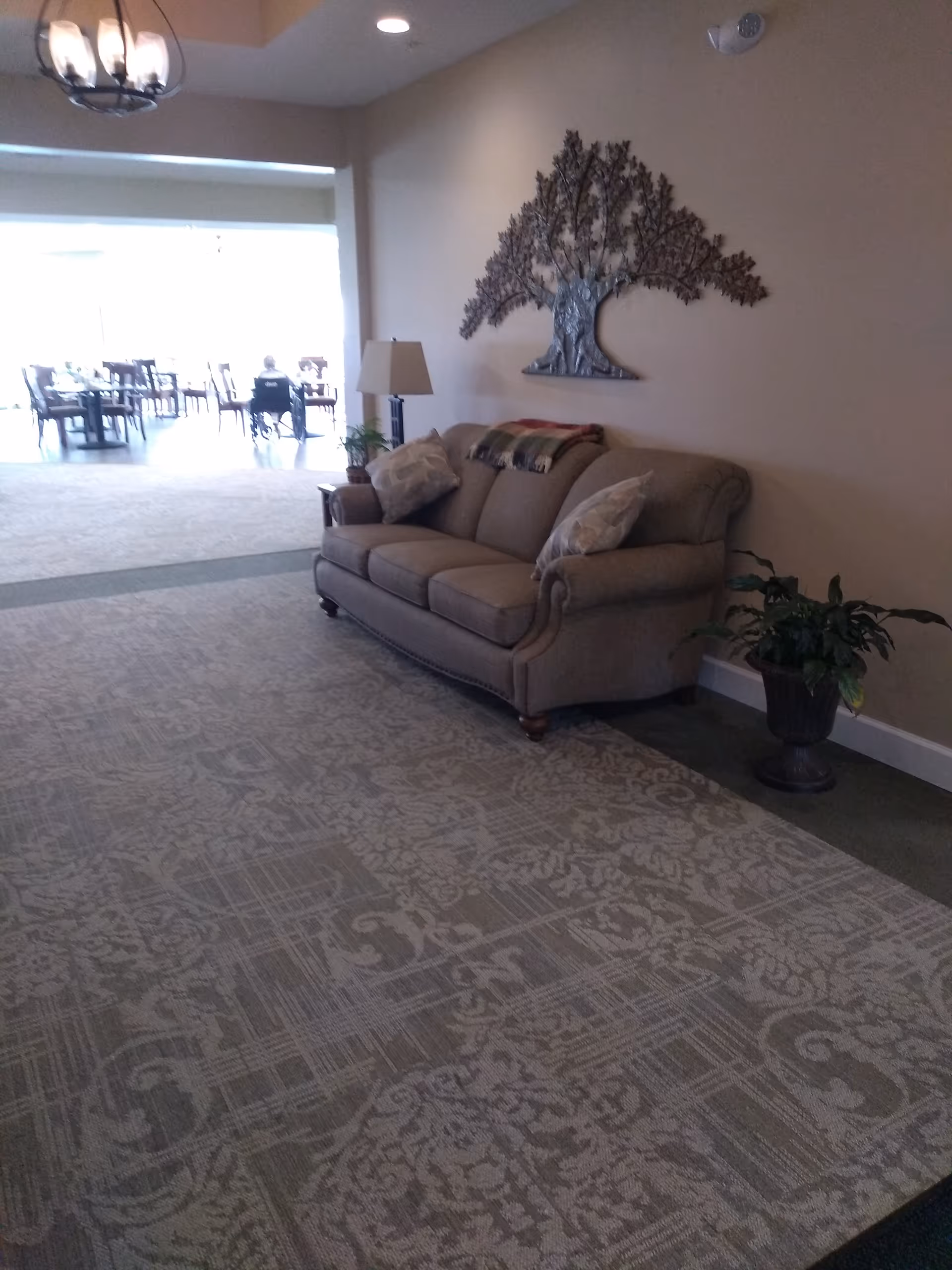 A cozy seating area in an assisted living facility with a beige sofa adorned with pillows and a blanket. A floor lamp and a potted plant are beside the sofa. On the wall above the sofa is a decorative metal tree artwork. In the background, there is a dining area with tables and chairs, and a person sitting in a wheelchair.