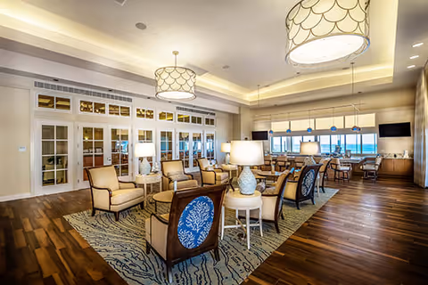 A spacious and well-lit living room area with multiple armchairs and small round tables arranged on patterned rugs. The room features large windows with a view of the ocean, modern ceiling lights, and wooden flooring. There are also built-in cabinets and a long counter with stools near the windows.