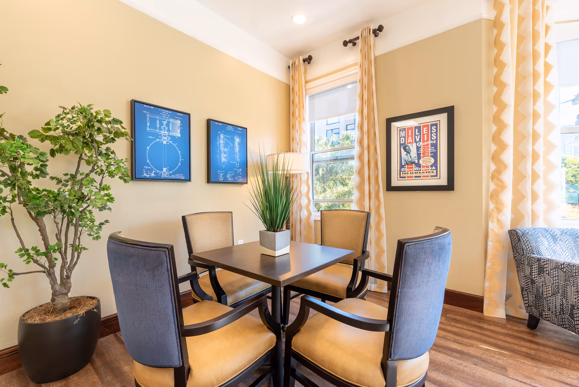 Small dining area with a square table surrounded by four upholstered chairs, a potted plant, framed artwork, and a window with patterned curtains.