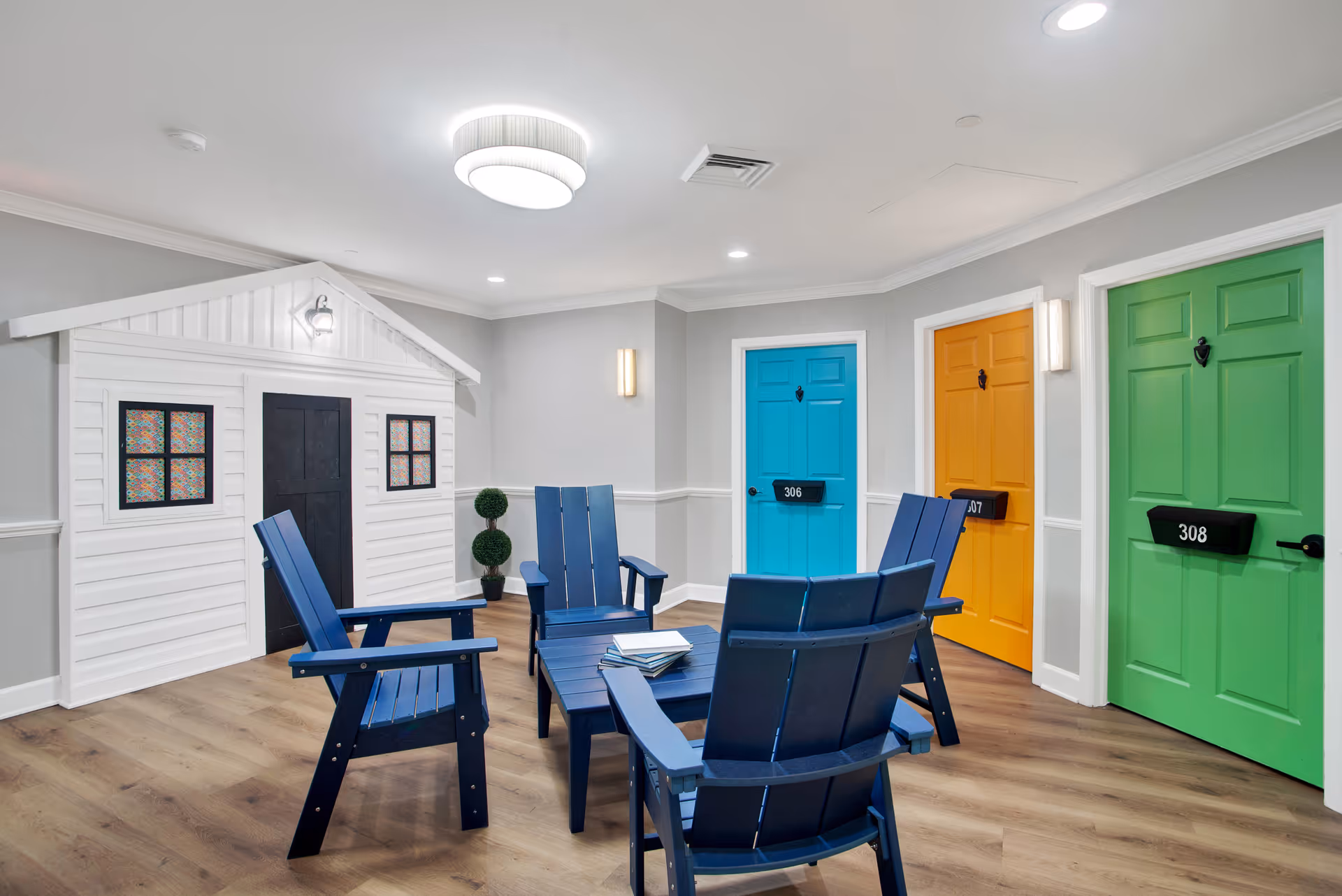 A bright and colorful indoor seating area with four blue wooden chairs arranged around a matching blue coffee table. Behind the chairs are three doors painted in vibrant colors: blue, orange, and green, each with a black mailbox and door knocker. To the left is a small white playhouse structure with a black door and two windows with colorful curtains. The room has light gray walls, wood flooring, and modern ceiling and wall lights.