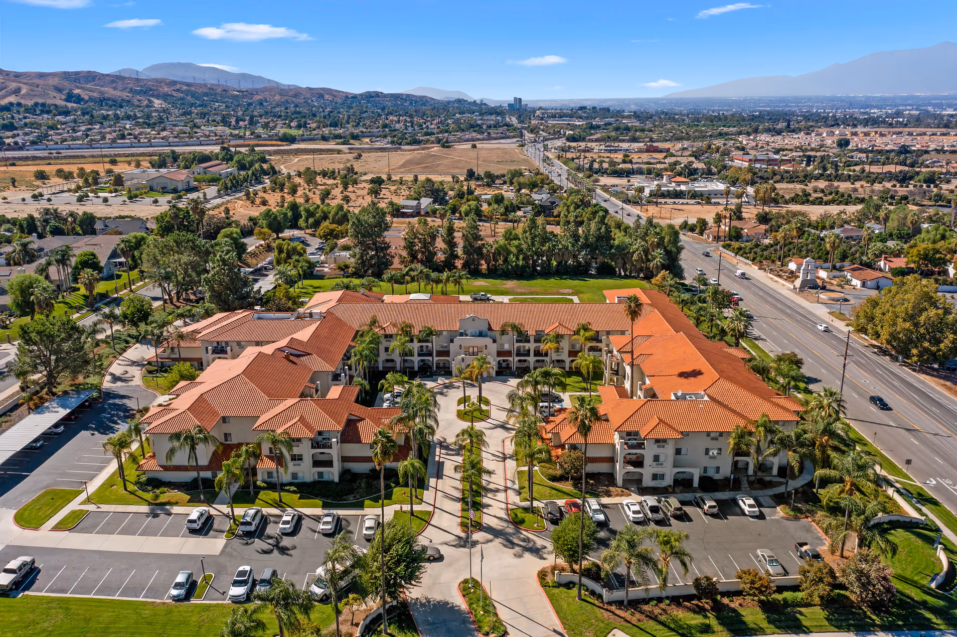 Aerial view of Mission Commons - A Provincial Senior Living Community, showing a large U-shaped building with red tile roofs surrounded by palm trees and parking lots. The facility is located in a suburban area with roads, houses, and hills in the background under a clear blue sky.
