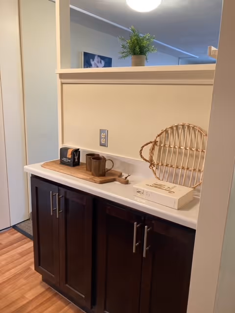 A countertop with dark wooden cabinets below, featuring a small wooden tray with a brown mug and a box, a wicker basket, and a small potted plant on the half wall above. The floor is wood, and a painting is partially visible on the wall in the background.