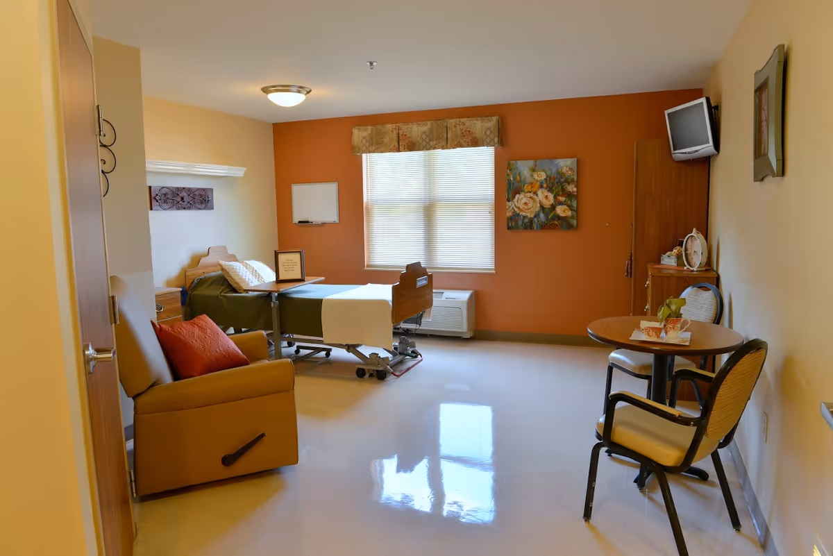 A well-lit nursing facility room with a hospital bed positioned near a window with blinds. The room features a comfortable recliner chair with a red pillow, a small round table with two chairs, a wall-mounted TV, and a wooden cabinet. The walls are painted in warm tones, with one accent wall in orange and the others in beige. There is a floral painting on the wall and a small whiteboard near the window.