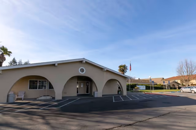 Exterior view of a single-story building with arched entrances and a parking lot in front. There are palm trees and an American flag near the building, with a clear blue sky overhead.