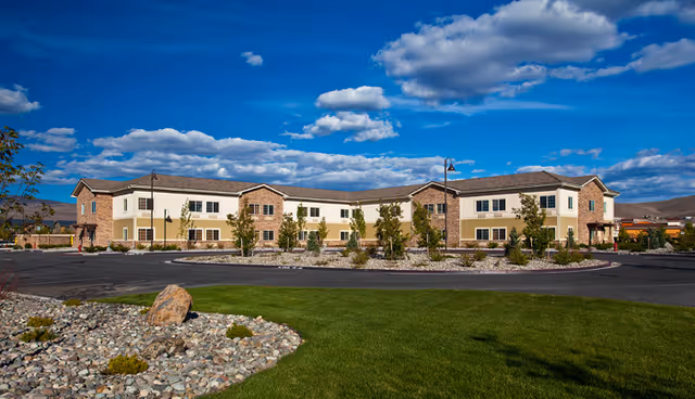 Exterior view of a two-story building with beige and brick facade under a blue sky with scattered clouds. The building is surrounded by a circular driveway, landscaped areas with rocks, shrubs, and green grass.