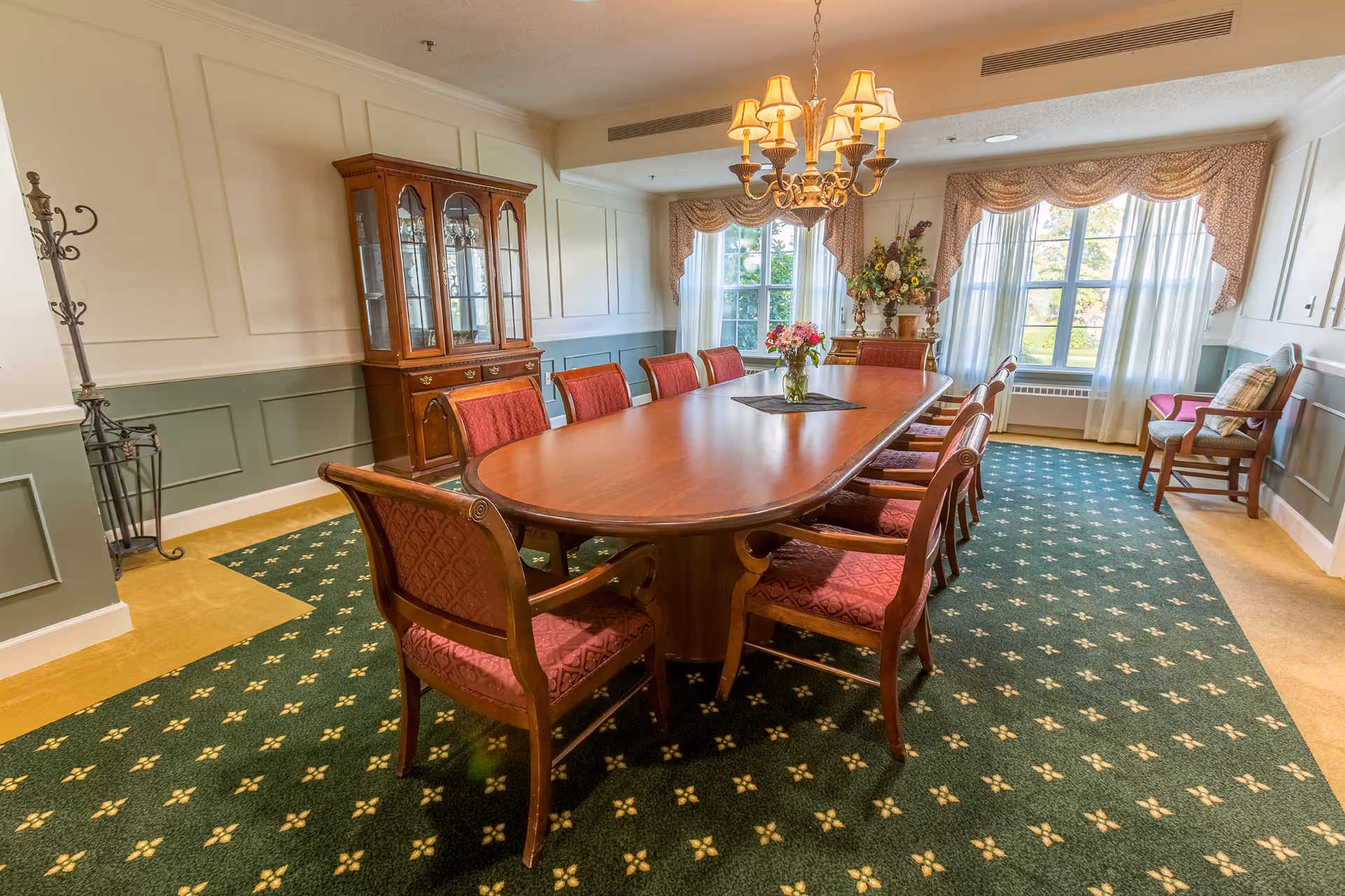 A formal dining room with a long wooden table surrounded by red upholstered chairs. A chandelier with multiple lamps hangs above the table. There is a vase with flowers on the table. The room has large windows with sheer curtains and decorative valances, a wooden china cabinet against one wall, and a green carpet with a yellow pattern.