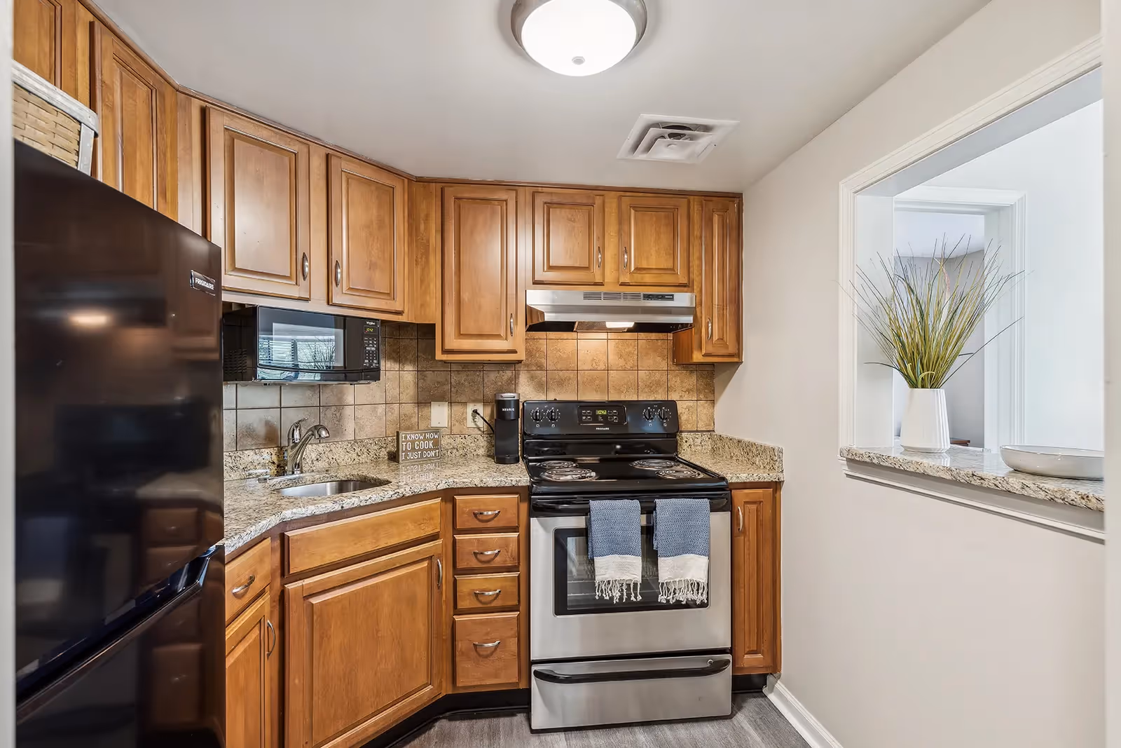 A compact kitchen with wooden cabinets, granite countertops, a black refrigerator, a stainless steel stove with two blue and white towels hanging on the handle, a microwave, and a small sink. There is a small decorative sign on the counter that reads 'I know how to cook I just don't'. A pass-through window with a vase of tall grass is visible on the right wall.
