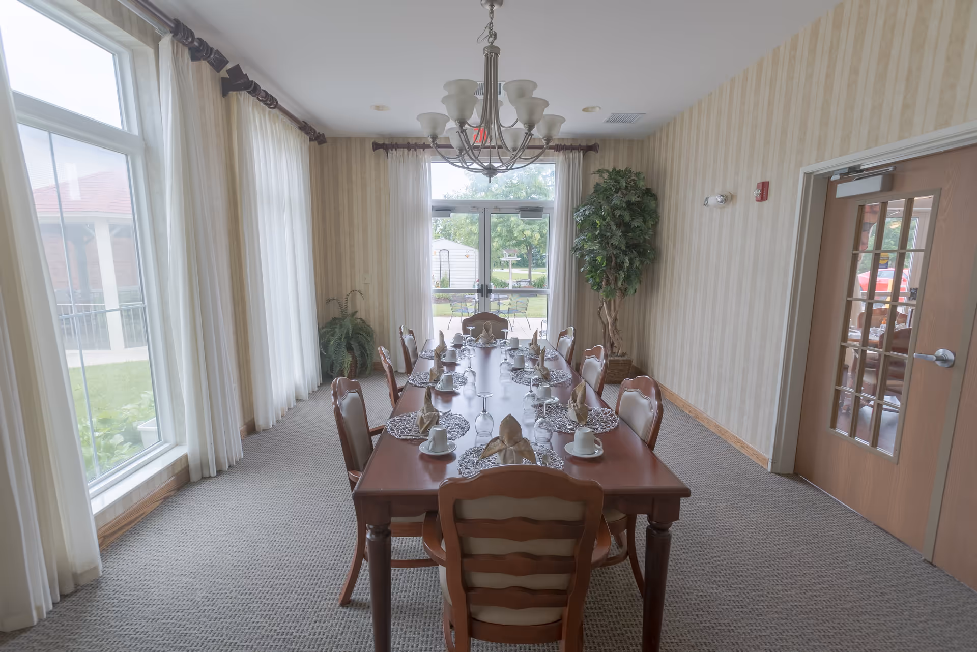 A long wooden dining table set for a meal in a bright dining room with large windows, a chandelier, and chairs.