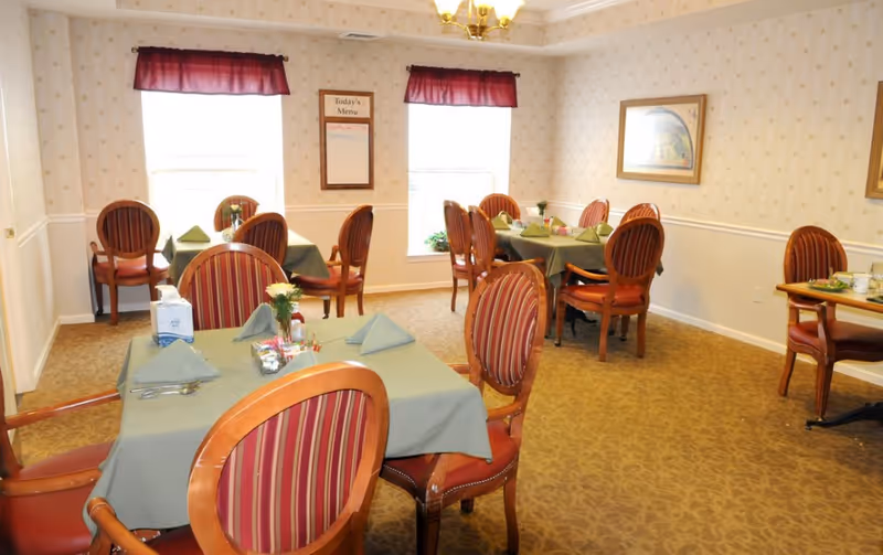 A dining room with several tables covered in green tablecloths, each set with folded napkins, utensils, and small flower arrangements. The chairs have wooden frames with red and gold striped upholstery. Two windows with maroon valances allow natural light into the room. A framed menu board and artwork hang on the wallpapered walls.