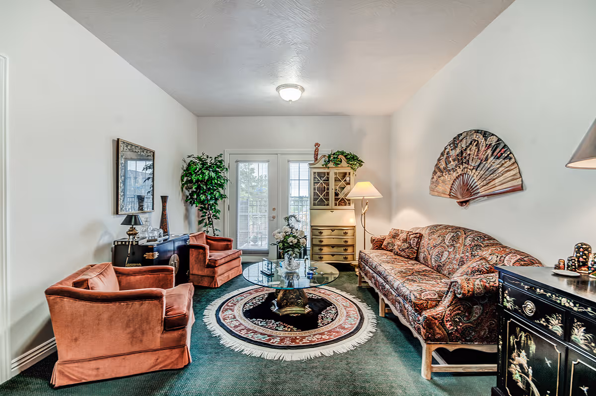 Well-lit living room featuring a patterned sofa, two rust-colored armchairs, a round glass coffee table on a circular rug, decorative cabinets, and French doors.