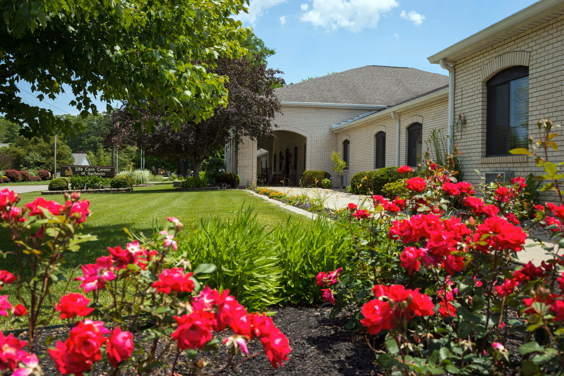 Exterior view of Life Care Center of Rochester showing a well-maintained garden with vibrant red flowers and green shrubs in the foreground, a manicured lawn, and a light-colored brick building with arched windows and an entrance under a clear blue sky.