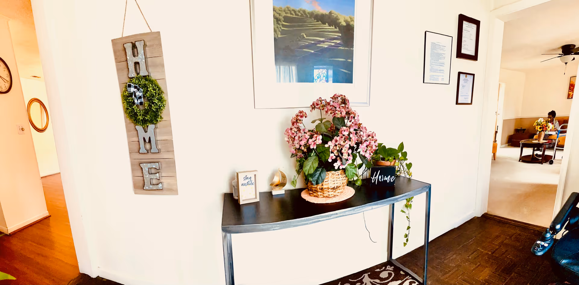 Interior view of a hallway corner in an assisted living facility. A narrow black table against a white wall holds a basket of pink flowers, a small plant, a decorative sailboat, and a sign that says 'Home'. Above the table is a framed picture. To the left, a wooden wall hanging spells out 'HOME' with a green wreath replacing the letter O. The floor is dark wood with a patterned rug underneath the table. In the background, a room with carpeted floor and a person sitting at a table with flowers is visible.
