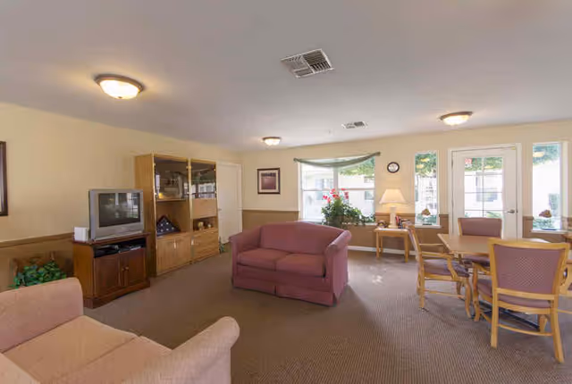 A senior living facility common area with beige walls and carpeted floor. The room features a pink loveseat and armchair, a wooden entertainment center with a TV, a wooden dining table with four chairs, and a side table with a lamp and plant. Large windows and glass doors allow natural light to enter the room.