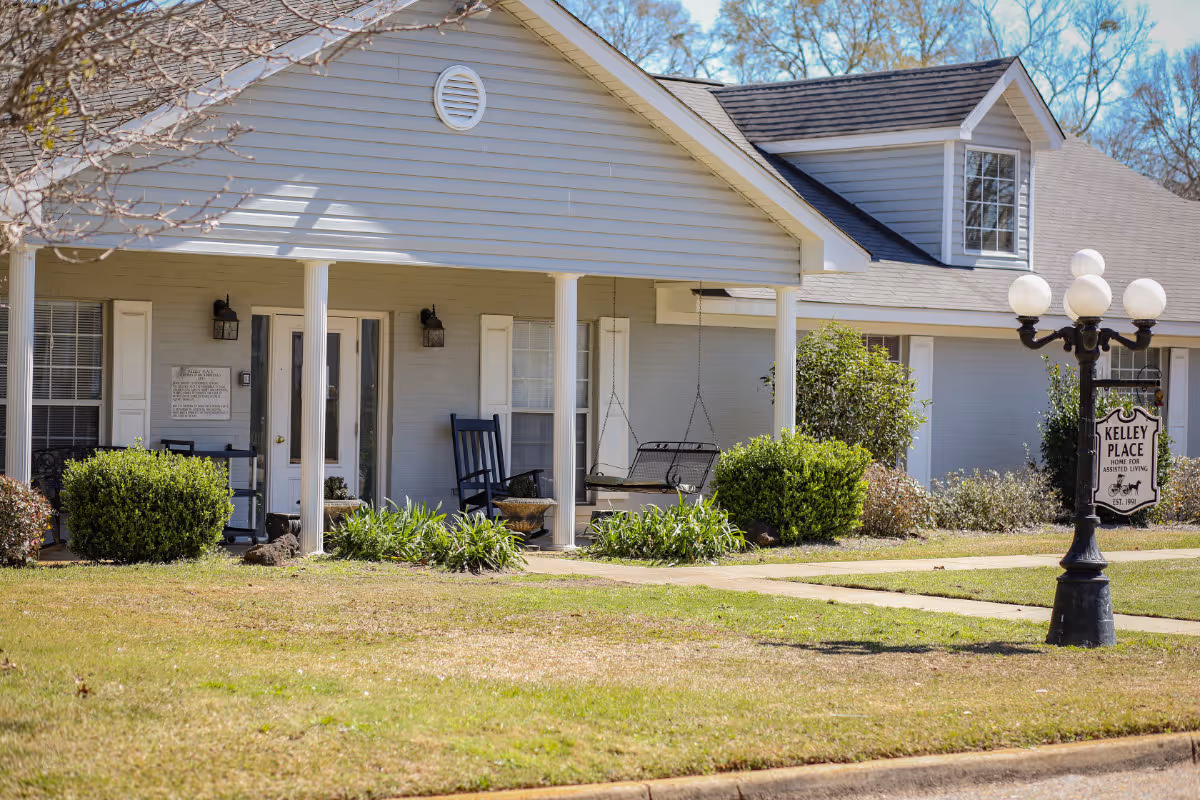 Exterior view of a single-story assisted living facility with a covered porch featuring white columns, rocking chairs, and a porch swing. The building has light gray siding and white trim, with bushes and plants in front. A vintage-style street lamp with a sign reading 'Kelley Place Home for Assisted Living Est. 1991' is visible on the lawn.