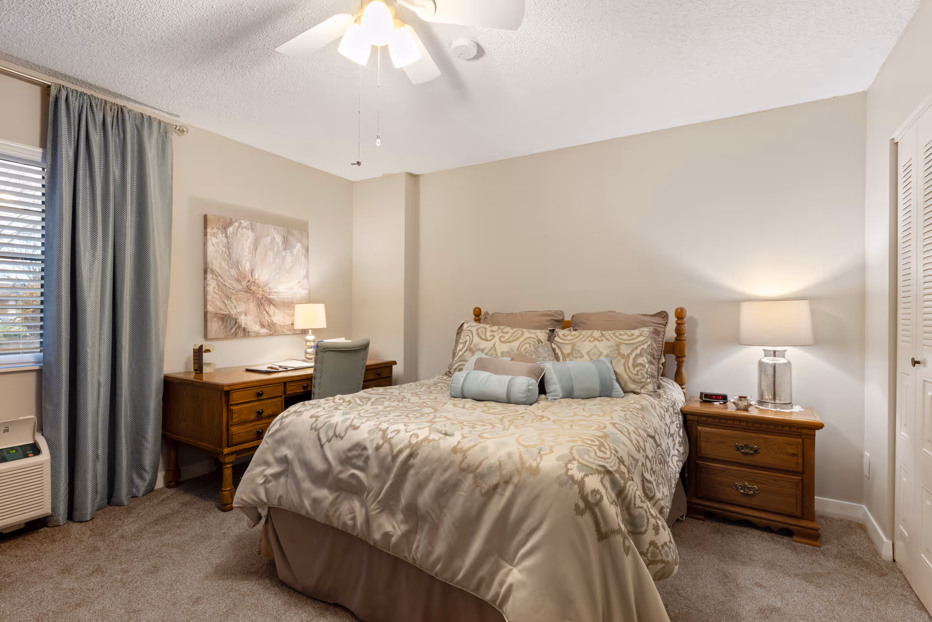 A cozy bedroom with a large bed covered in a patterned beige and light blue comforter and multiple pillows. There is a wooden nightstand with a lamp and an alarm clock on the right side of the bed. On the left side, there is a wooden desk with a chair, a lamp, and some items on top. A window with blue curtains and blinds is visible on the left wall. The room has beige walls and carpeted floor.