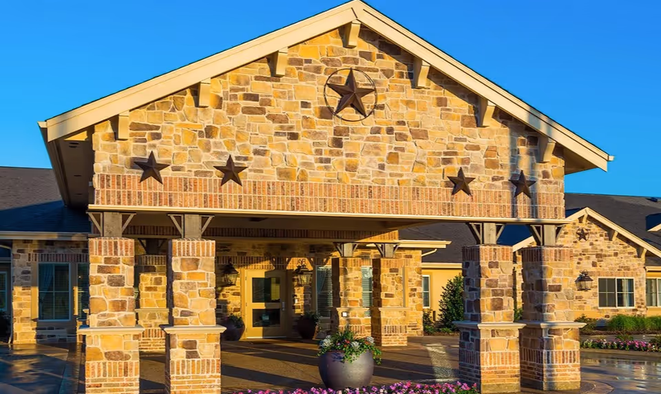 Exterior view of a senior living facility entrance with stone and brick pillars supporting a covered porch. The building features decorative star accents on the stone facade and a clear blue sky in the background. There are potted plants and flowers near the entrance.