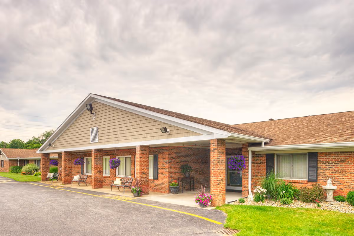 Exterior view of a single-story brick building with a covered entrance supported by brick columns. There are benches and hanging flower baskets under the covered area, with a well-maintained lawn and garden beds nearby. The sky is cloudy.