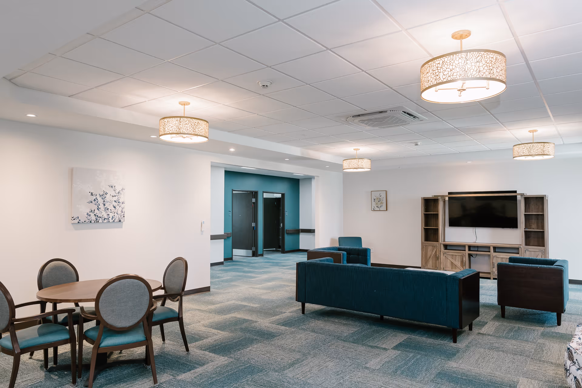 A spacious senior living common area with teal carpet and white walls. The room features a round wooden table with four chairs on the left, and a seating area with a teal sofa, two armchairs, and a wooden entertainment center with a flat-screen TV on the right. The ceiling has multiple decorative light fixtures, and there are two doorways visible in the background.