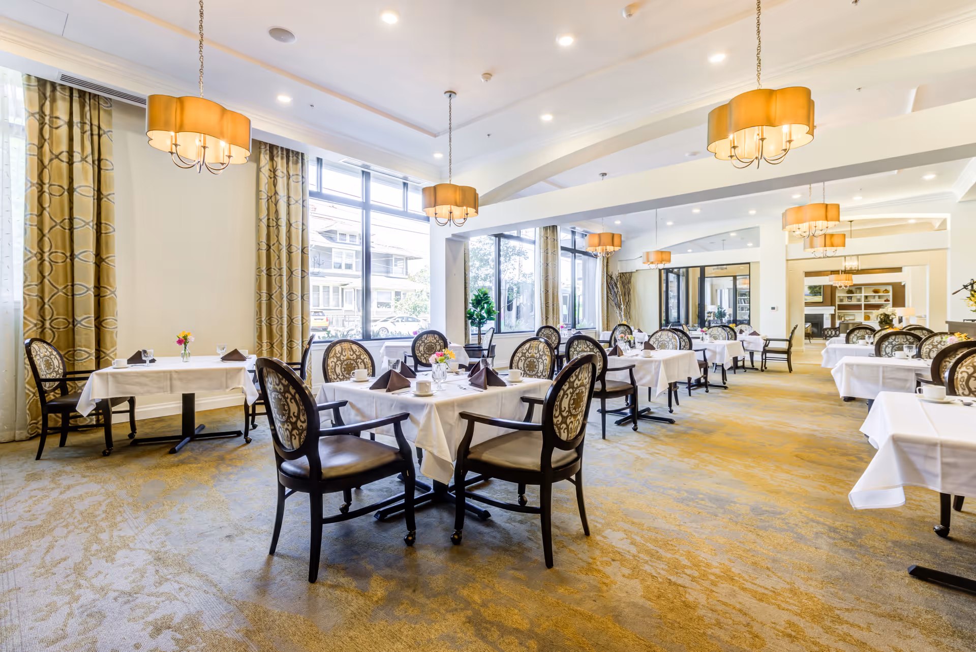 A bright and spacious dining room with multiple tables covered in white tablecloths, each set with cups, glasses, napkins, and small flower arrangements. The room features large windows with patterned curtains, elegant upholstered chairs, and warm pendant lighting hanging from the ceiling.