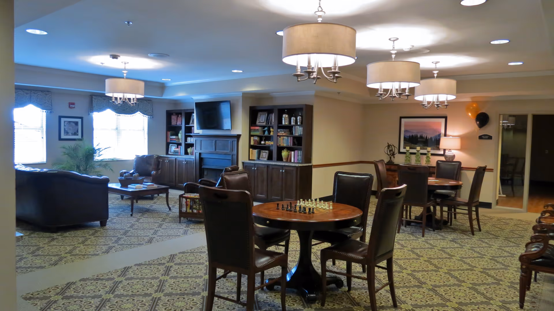 A cozy senior living community common area with patterned carpet, round wooden tables with chairs, one table set up with a chessboard, leather armchairs, a fireplace with built-in bookshelves, a wall-mounted TV, and soft overhead lighting from chandeliers.