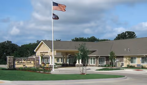 Exterior view of Hudson Creek Alzheimer's Special Care Center building with a flagpole flying the American flag and another flag in front. The building is single-story with a beige facade and a covered entrance. There is a landscaped area with grass, trees, and flowers near the entrance and a sign displaying the facility's name.