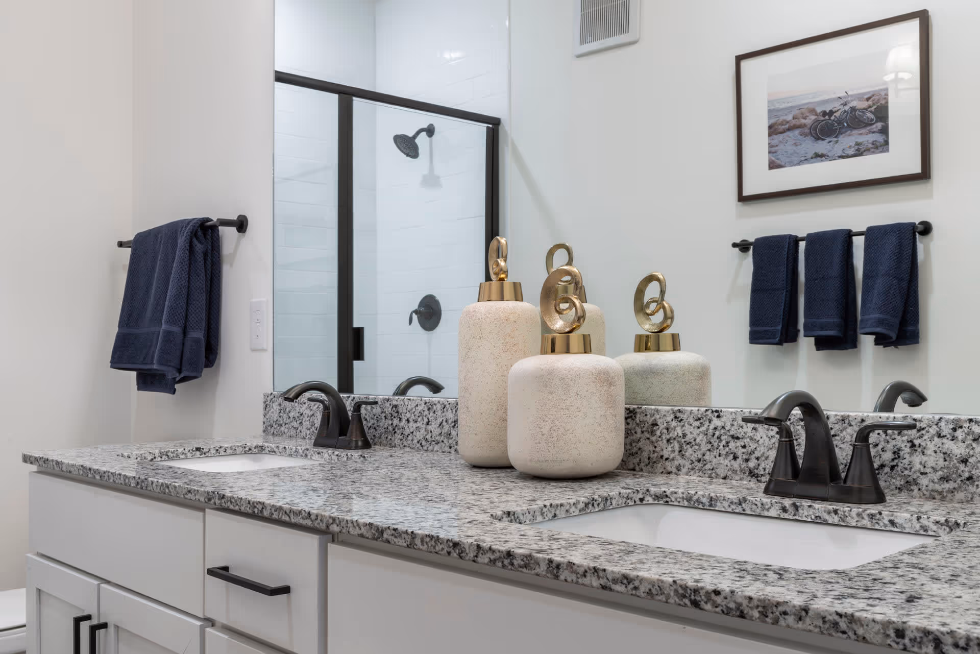 Modern bathroom with a granite countertop featuring two sinks and black faucets. There are decorative vases on the counter, a large mirror above, and a glass-enclosed shower with a black showerhead in the background. Dark blue towels hang on black towel racks on the walls, and a framed picture is visible above one towel rack.