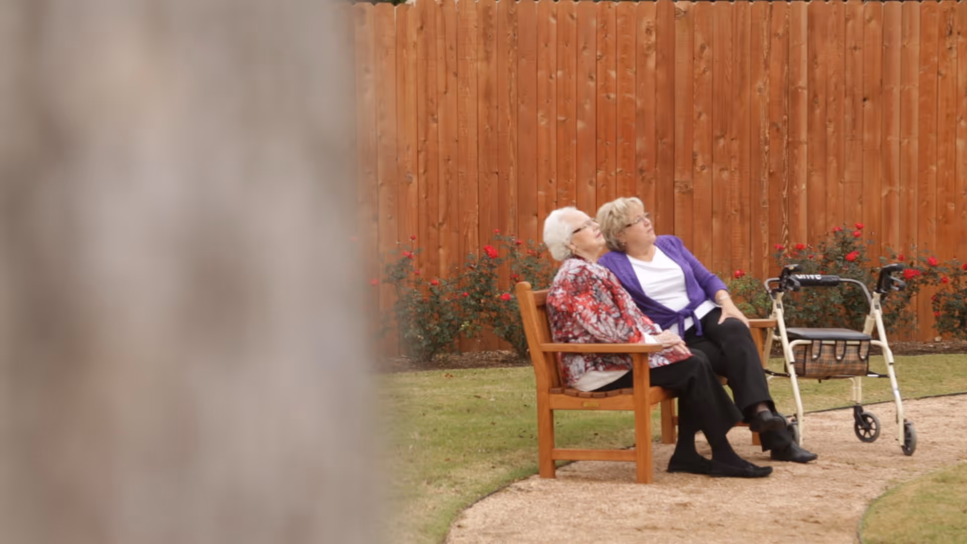 Two elderly women sitting on a wooden bench in a garden area with a wooden fence and rose bushes in the background. One woman is wearing a red floral jacket and the other a purple cardigan. A walker is positioned next to the bench.