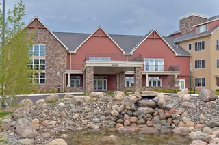 Entrance façade of a red and stone multi-story senior living building with a rock-lined pond and small waterfall in the foreground.