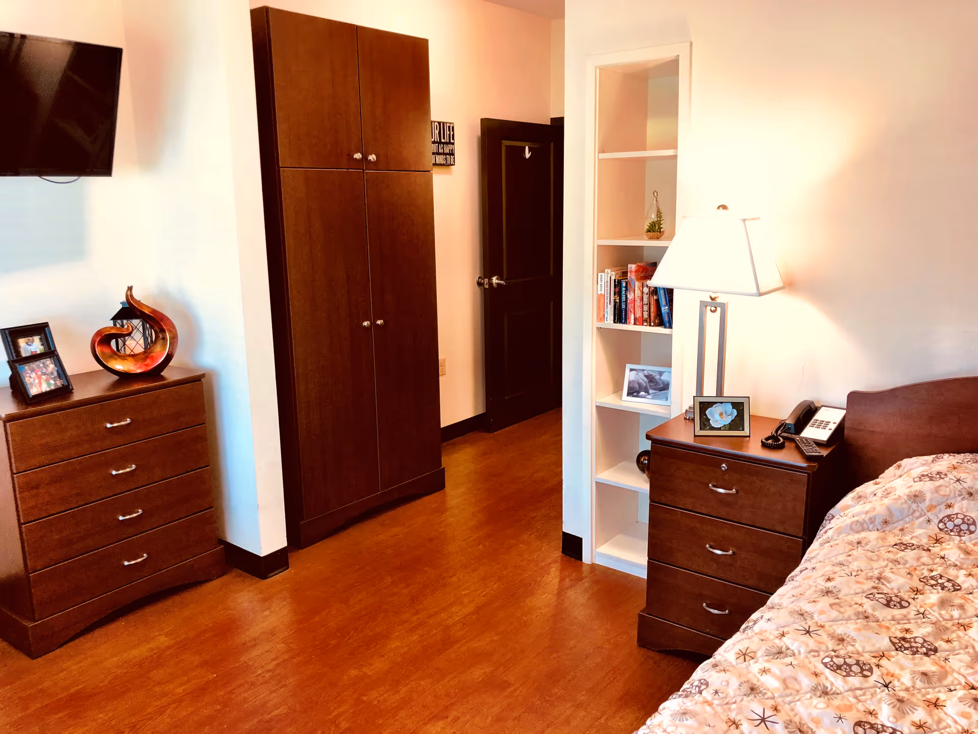 A furnished bedroom with wooden dressers, built-in shelving, a bedside lamp and phone, wall-mounted TV, and a bed with patterned bedding.
