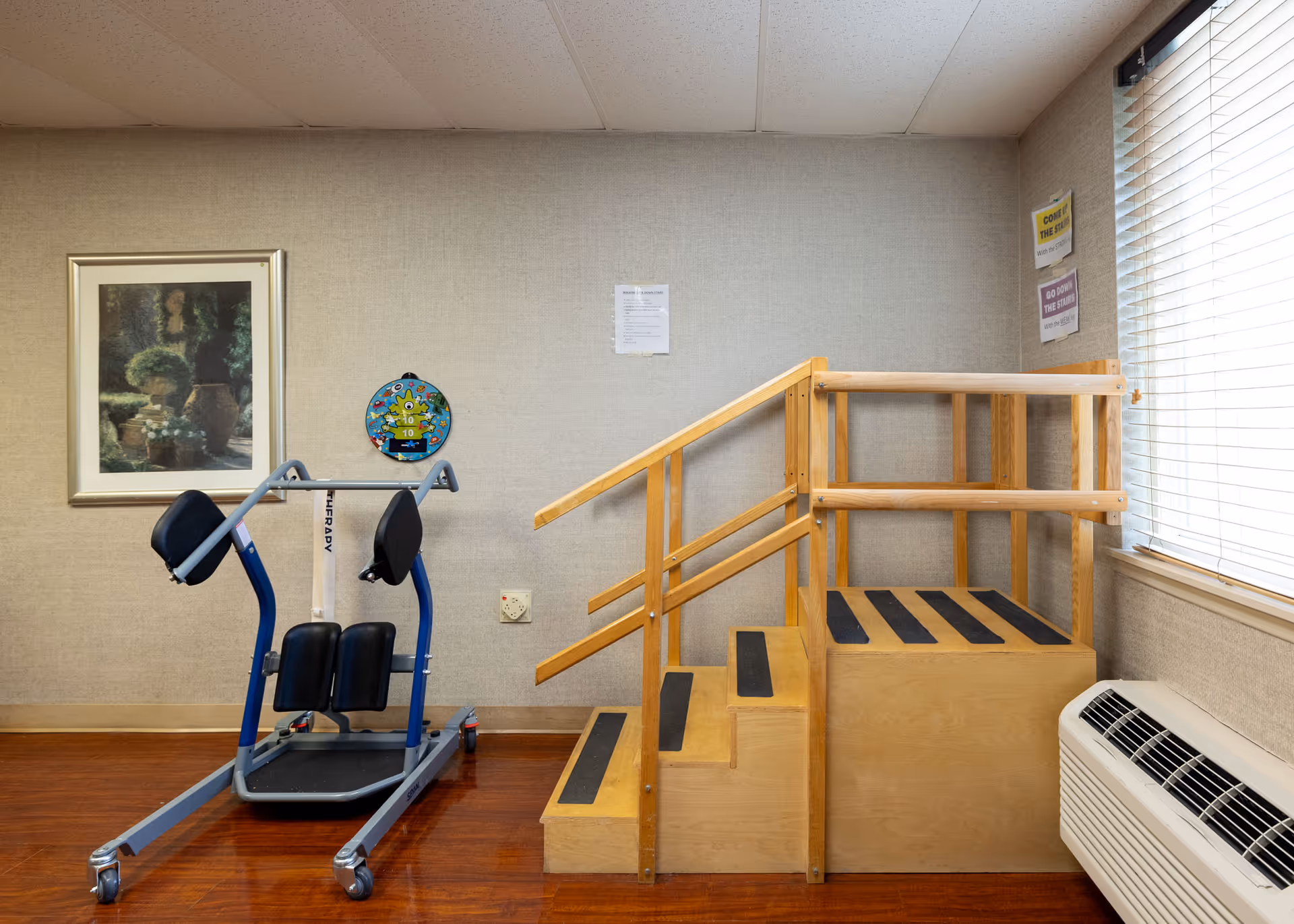 A therapy room with a wooden staircase with handrails and a blue exercise machine on wheels. The room has beige walls, a framed picture of a garden scene, a colorful target on the wall, and a window with blinds. There is also a white air conditioning unit below the window.