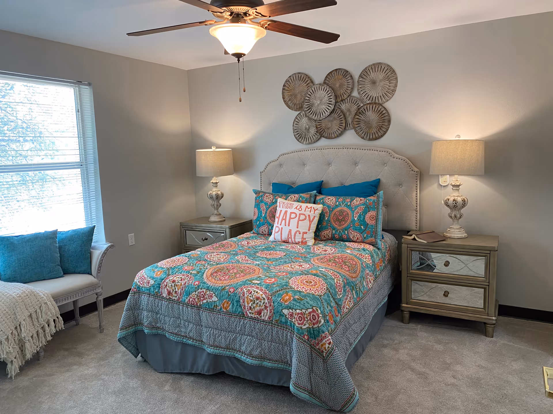 A cozy bedroom with a bed featuring a colorful patterned quilt and multiple pillows, including one with the text 'This is my happy place.' The bed has a tufted headboard and is flanked by two matching nightstands with lamps. A ceiling fan with a light is above the bed, and a window with blinds and a cushioned bench with blue pillows is on the left side of the room. Decorative wall art hangs above the headboard.