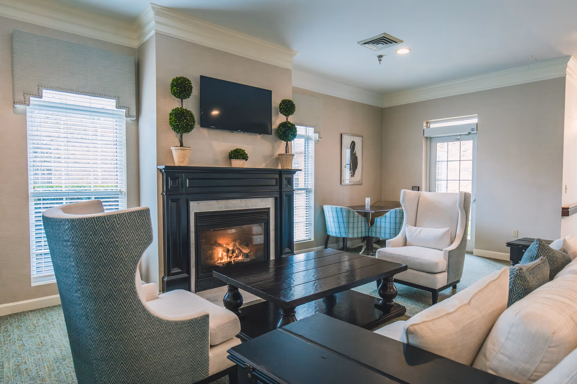A cozy living room area with a lit fireplace, a mounted flat-screen TV above it, two large windows with blinds on either side, two beige armchairs, a dark wooden coffee table, a beige sofa with cushions, and a small dining table with two blue plaid chairs near a glass door.