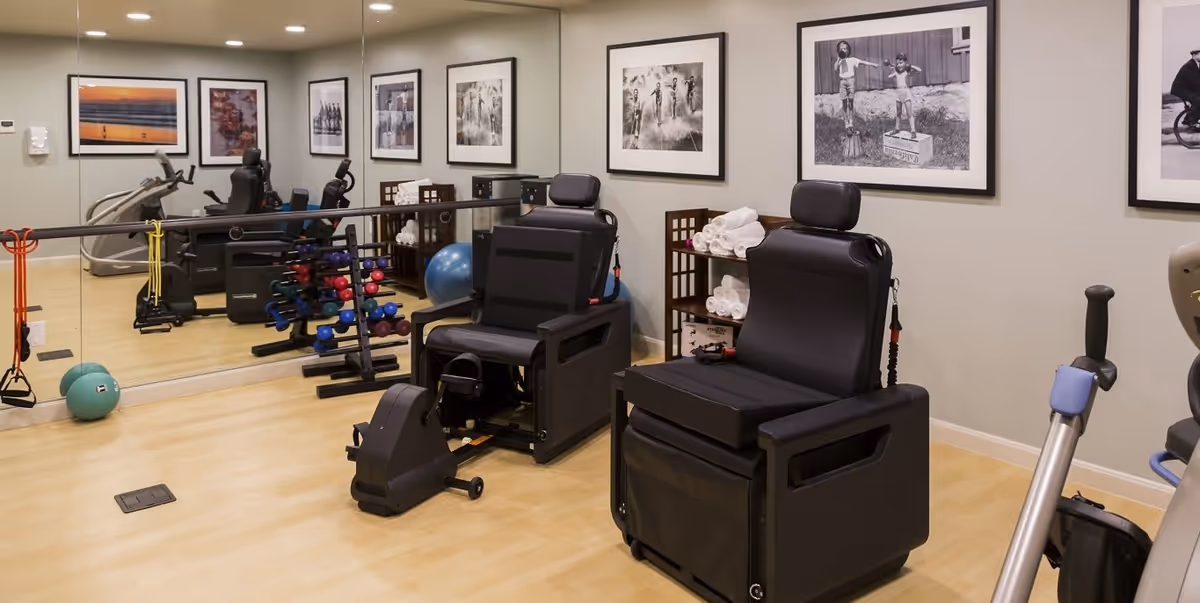 A fitness room with exercise equipment including two black exercise chairs, a stationary bike, a rack of colorful dumbbells, resistance bands, a blue exercise ball, and a large mirror on one wall. The walls are decorated with framed black and white photographs.