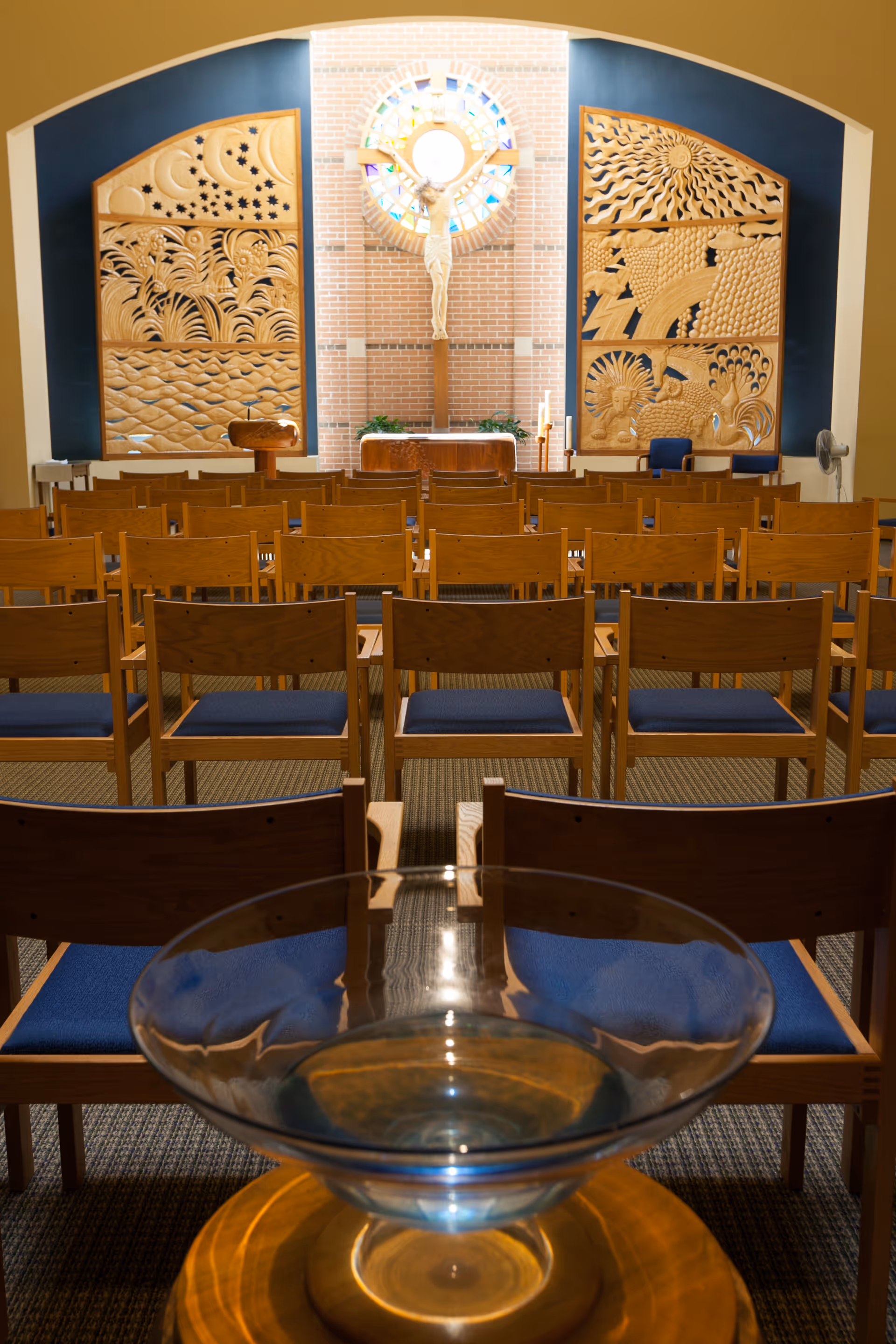 Interior view of a chapel with rows of wooden chairs with blue cushions facing an altar. Behind the altar is a crucifix mounted on a brick wall with a circular stained glass window above it. On either side of the altar are large wooden panels with carved religious and natural motifs.