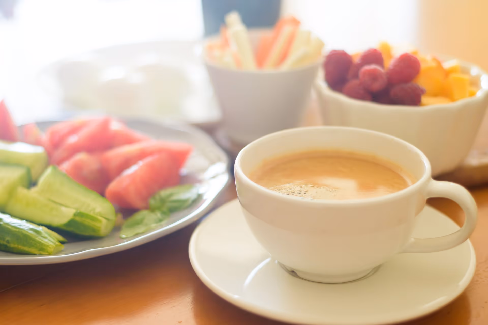 A white coffee cup on a saucer sits on a table with bowls of fruit in a bright dining setting.