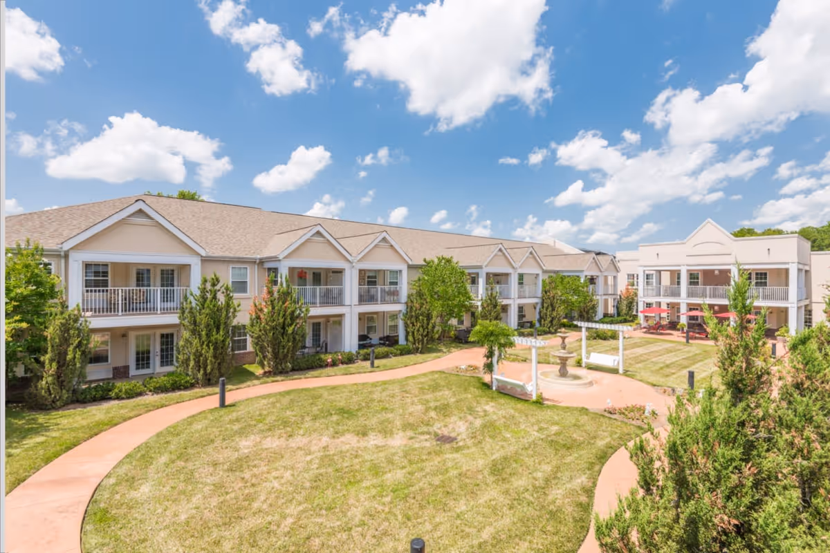 Two-story senior living building surrounding a landscaped courtyard with walkways and a central fountain under a blue sky.