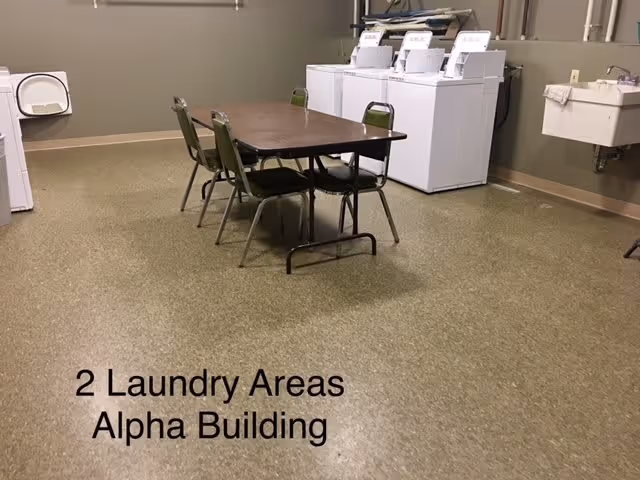 A laundry room with a row of washing machines and dryers against the wall, a table with four chairs in the center, and a utility sink on the right side. The floor is covered with speckled linoleum, and the walls are painted beige. Text on the floor reads '2 Laundry Areas Alpha Building'.