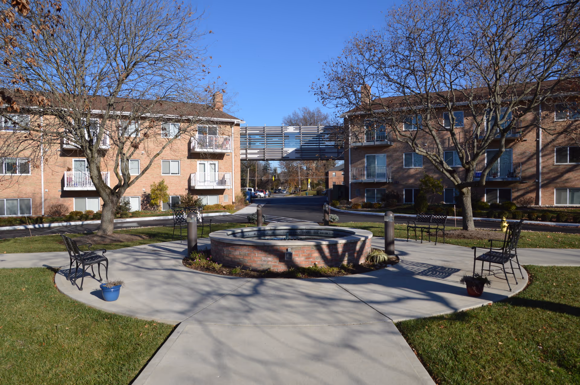 Courtyard with a circular brick fountain, benches, and walkways between two brick apartment buildings under a clear blue sky.
